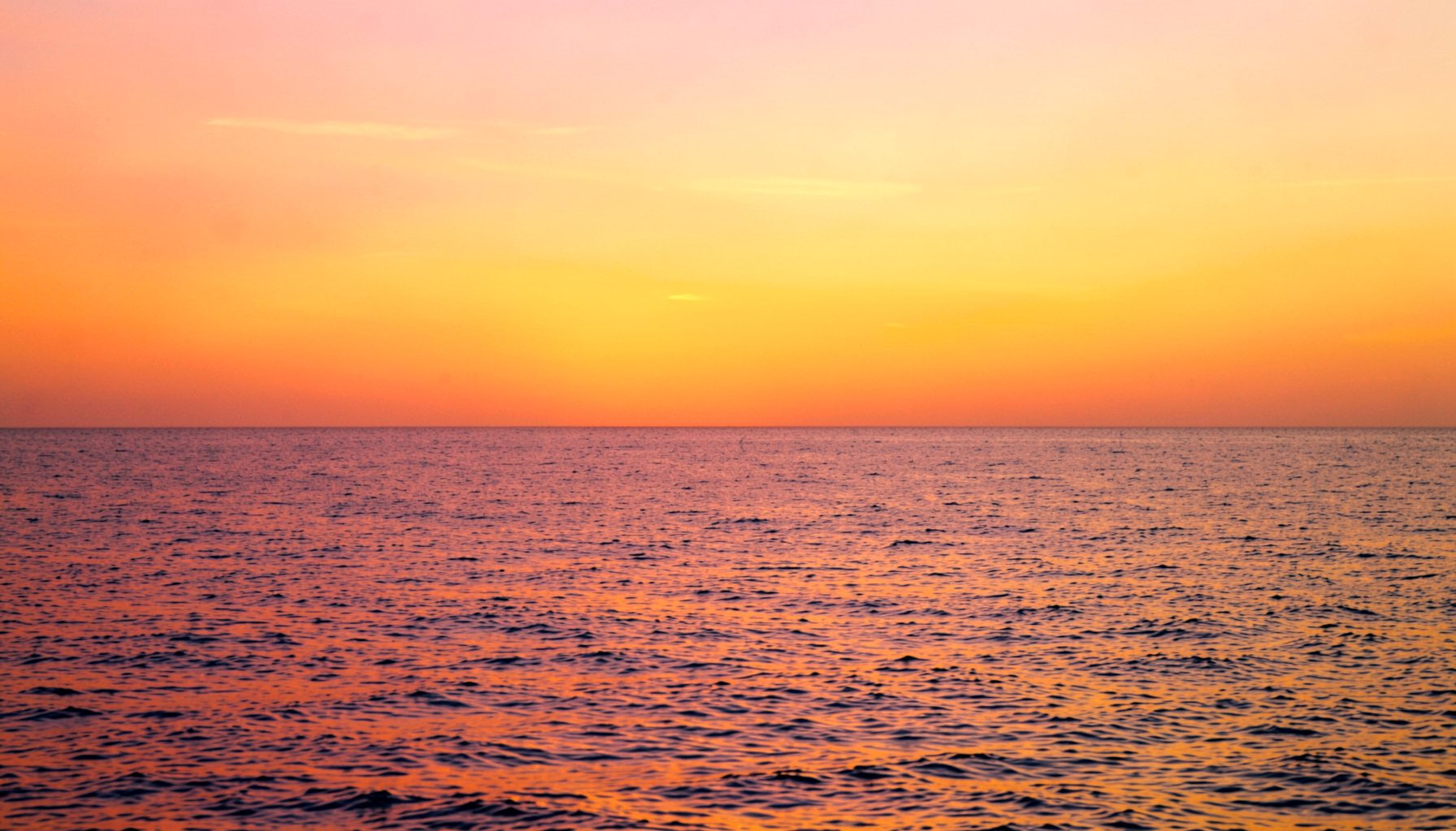 Warm sunset light over calm ocean water, blending deep orange, soft pink, and golden yellow across the horizon, with a tranquil and serene mood.